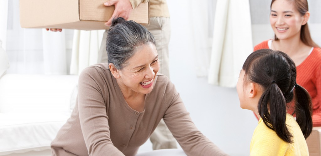 A woman playing with a child on the floor while a man carries a cardboard box in the background