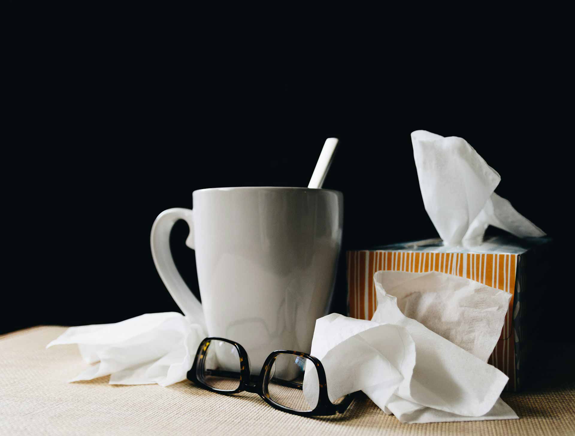 A mug of tea and a pair of glasses, sitting on a table with some crumpled up tissues